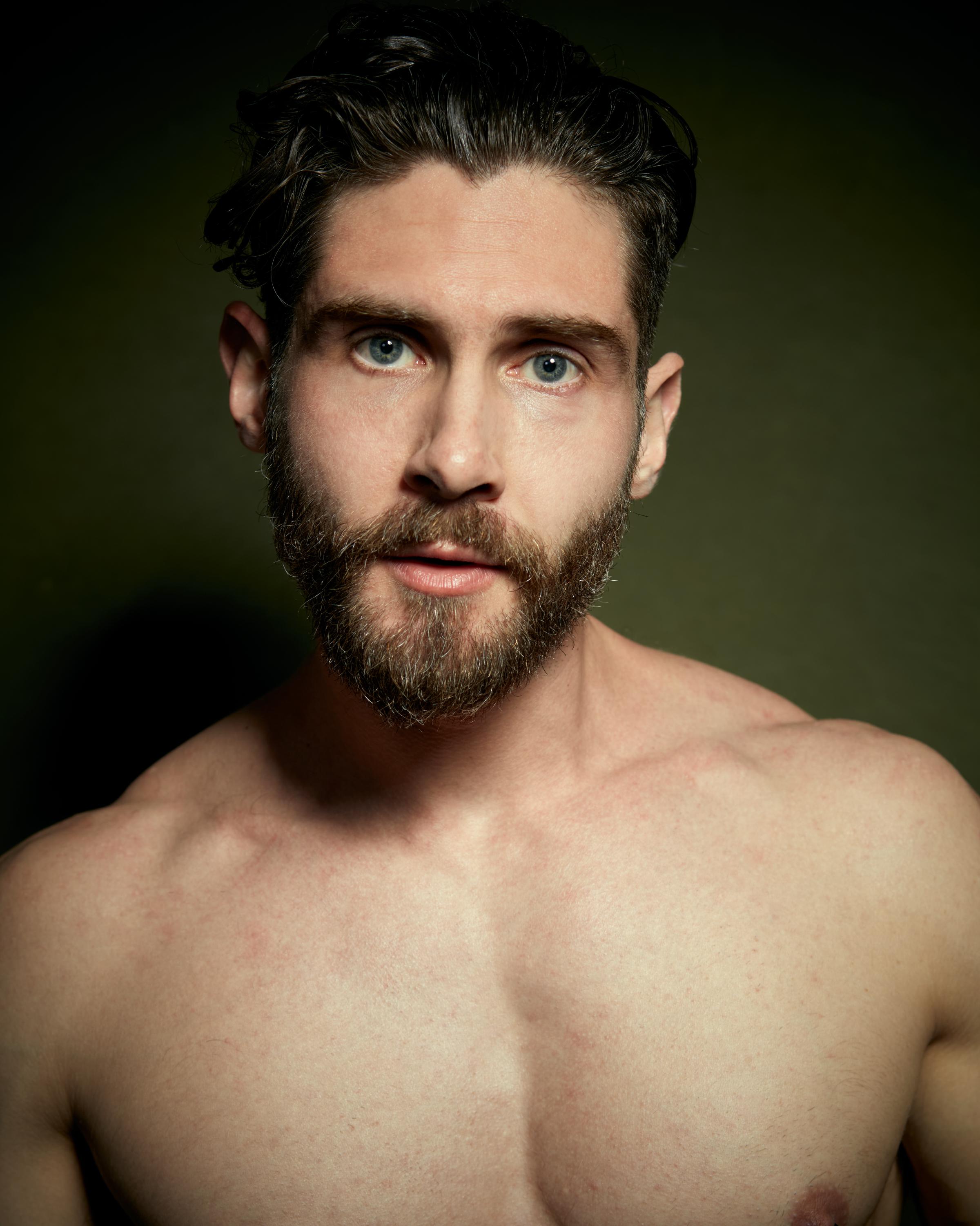 Close-up portrait of a bearded man with slicked-back hair and blue eyes, shirtless under dramatic light against an olive-toned backdrop.