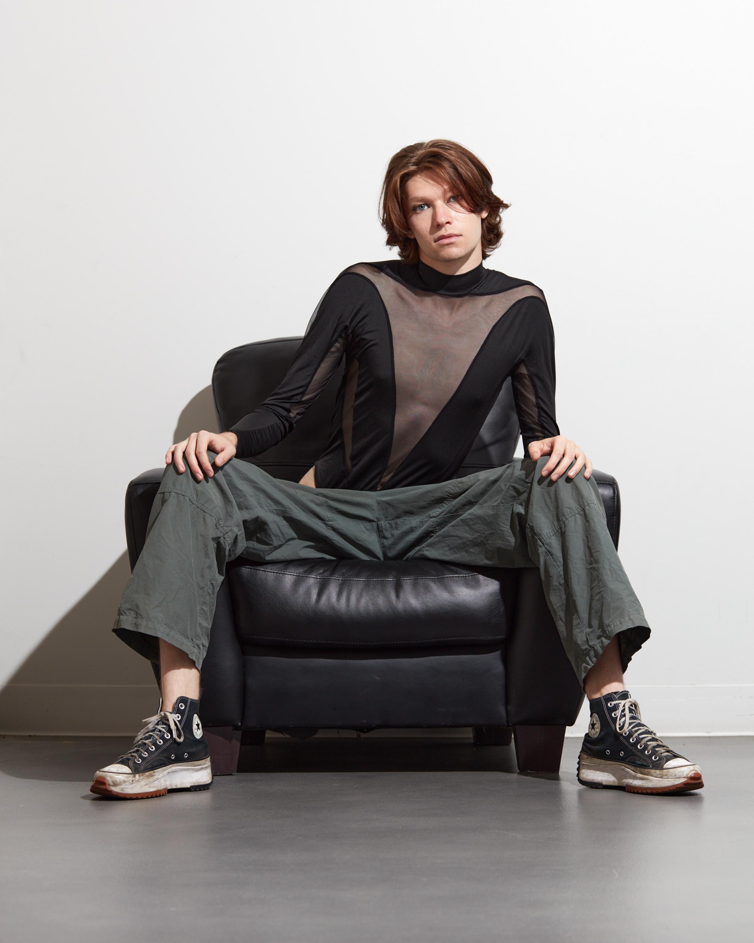 High-contrast studio fashion portrait of a male model in a sheer black top and wide-leg pants, seated on a leather chair.