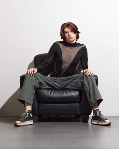 High-contrast studio fashion portrait of a male model in a sheer black top and wide-leg pants, seated on a leather chair.