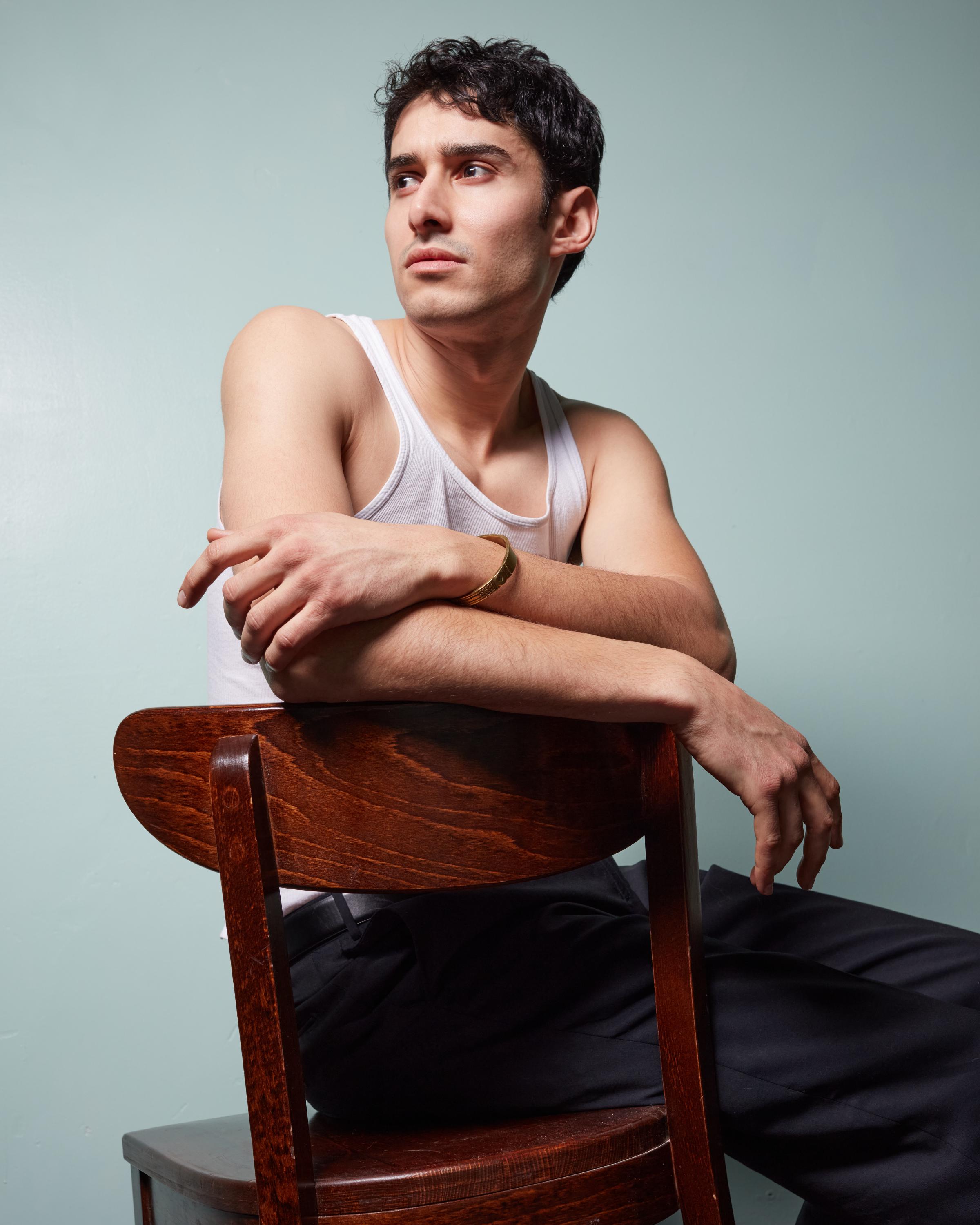 Man with short dark hair sitting backwards on a wooden chair, wearing a white tank top and black pants, looking thoughtfully to the side against a teal background.