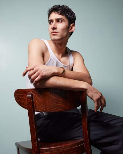 Man with short dark hair sitting backwards on a wooden chair, wearing a white tank top and black pants, looking thoughtfully to the side against a teal background.