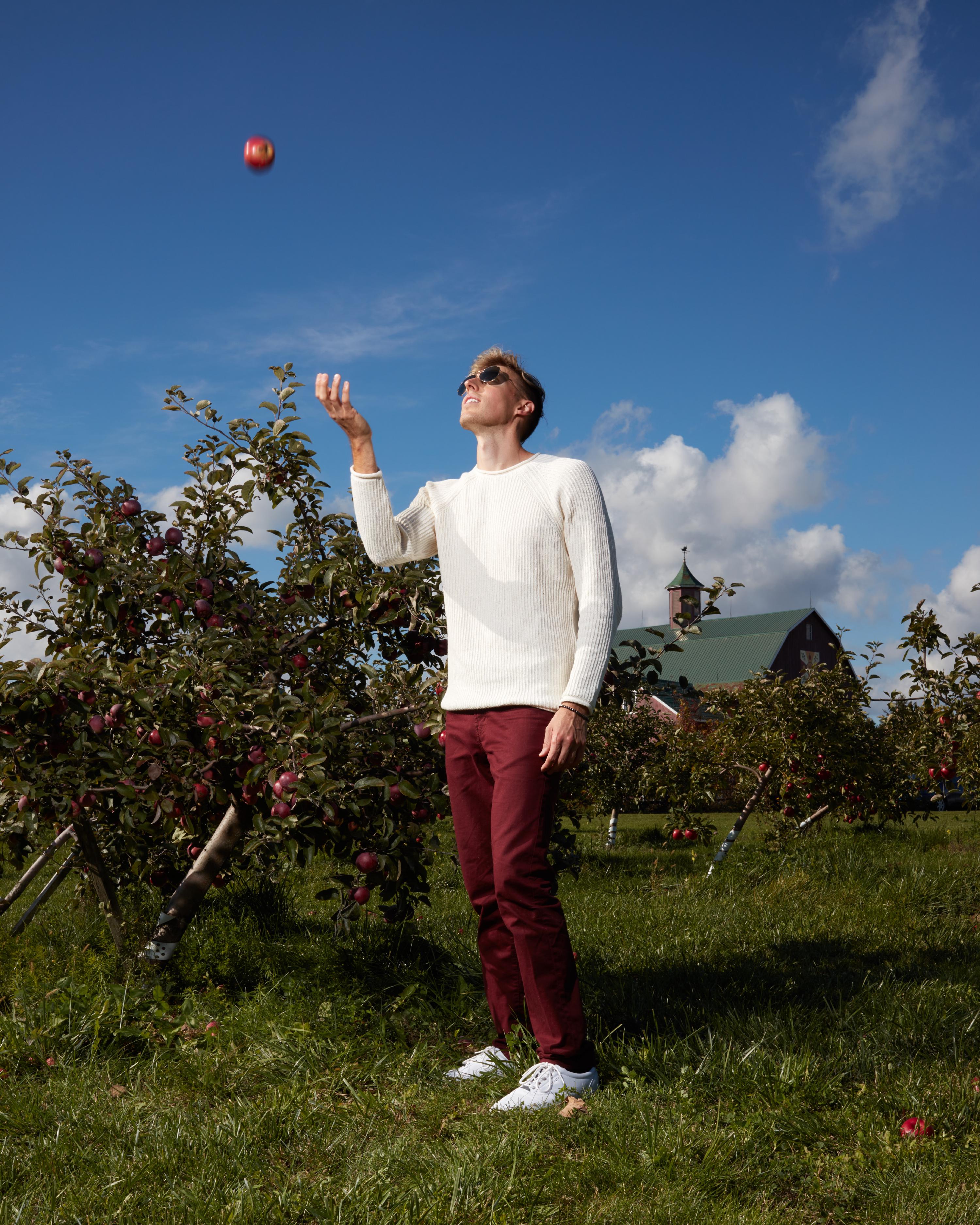 Man in white sweater juggling an apple in an orchard with apple trees and a barn under a clear blue sky.