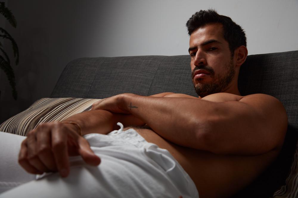 Man with short dark hair and a beard reclining shirtless on a gray sofa, looking directly at the camera with an intense and smoldering expression.