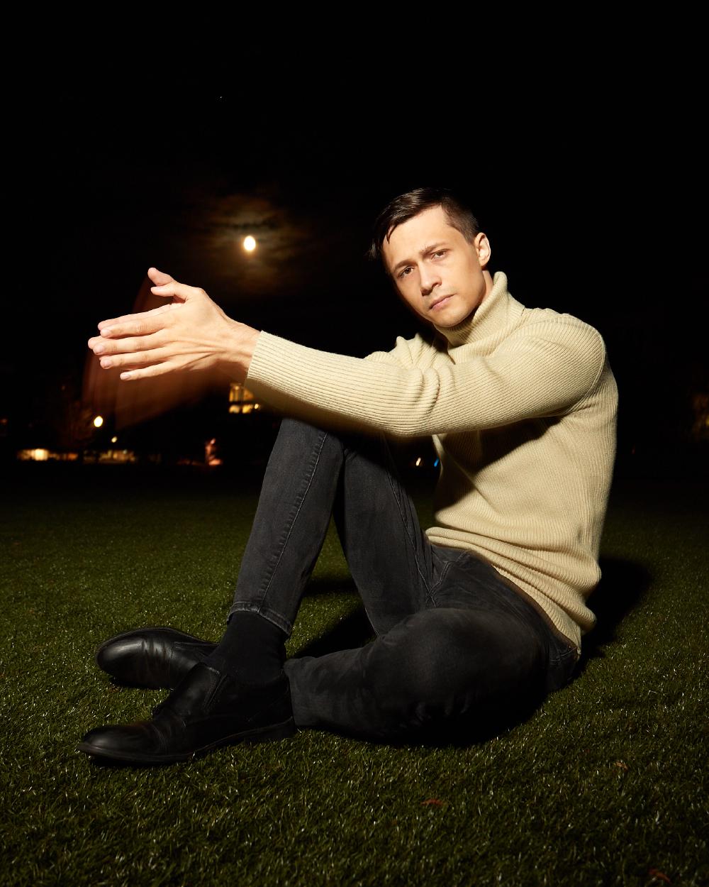 Man in a ribbed beige turtleneck sweater sitting on Tindall Field on the grass at night, extending his arms with a focused expression, under a dark sky illuminated by the moon.