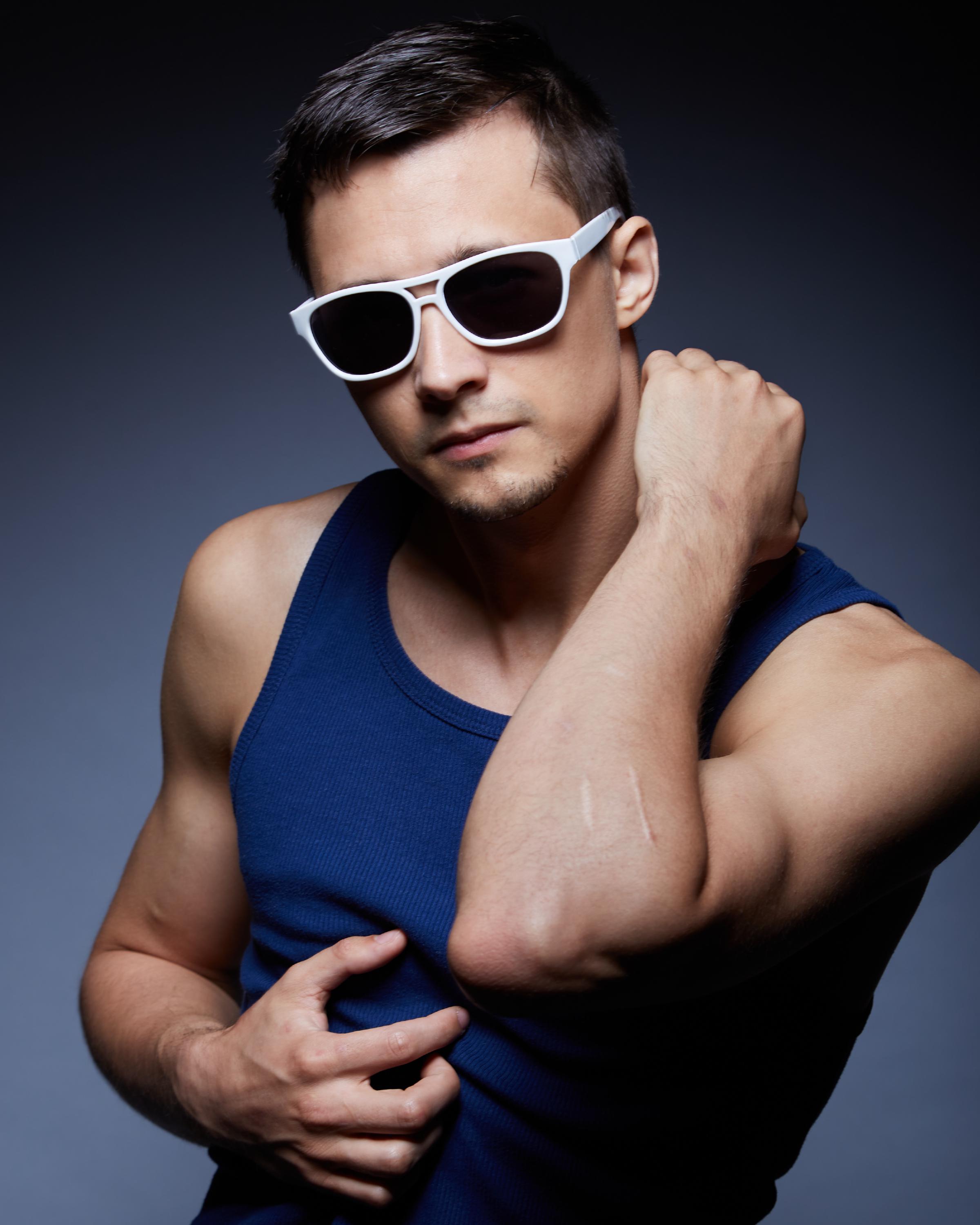 Man in a navy blue Aussiebum tank top and white sunglasses posing confidently with a stylish, intense expression.