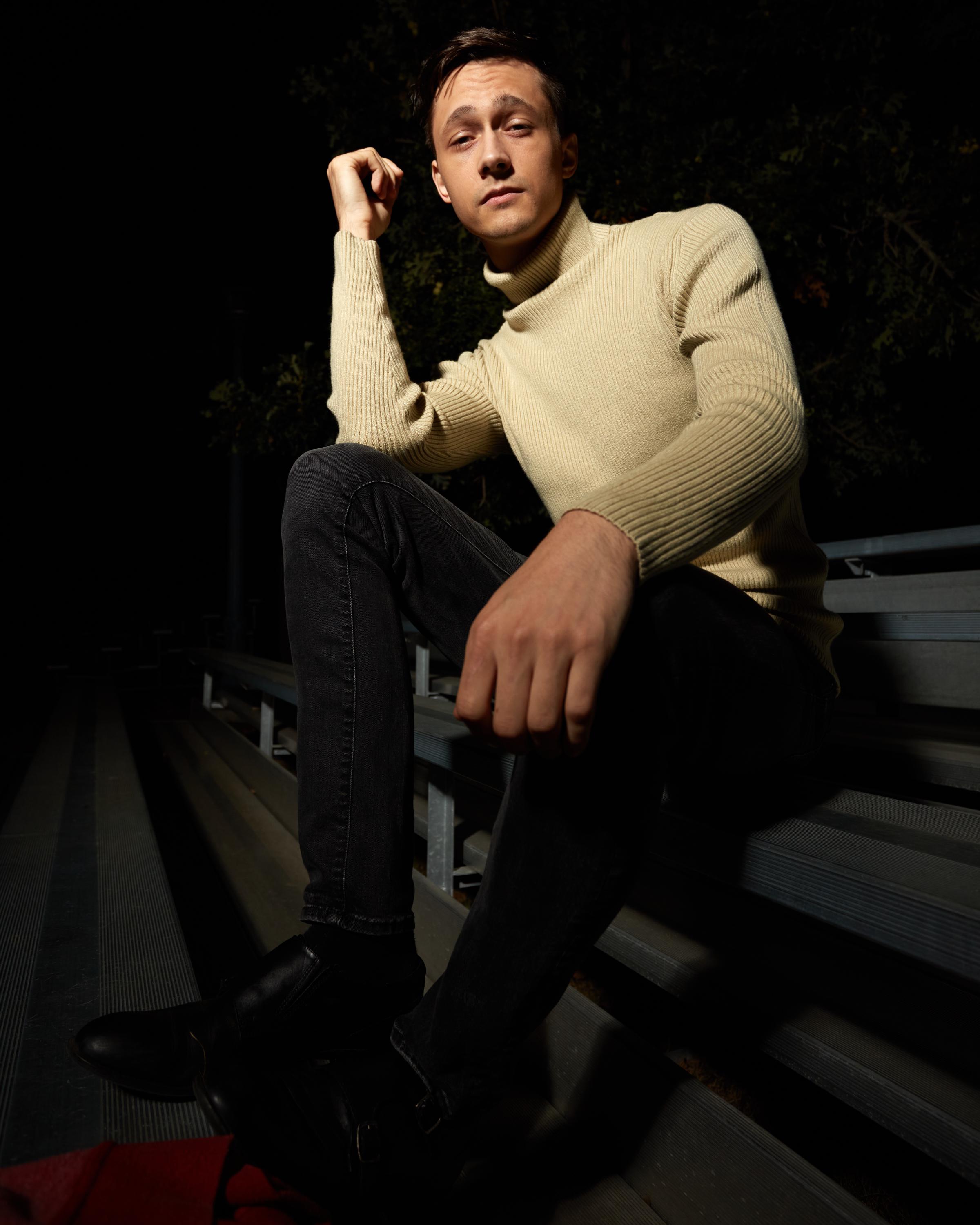 Low-angle editorial portrait of a man in a beige turtleneck sweater and black jeans sitting on the Tindall Field bleachers at Queen's University at night, with cinematic directional lighting and dramatic shadows.