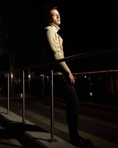 Man in a beige ribbed turtleneck sweater posing dramatically in front of the Isabel Bader Centre at night, with an intense and moody expression.