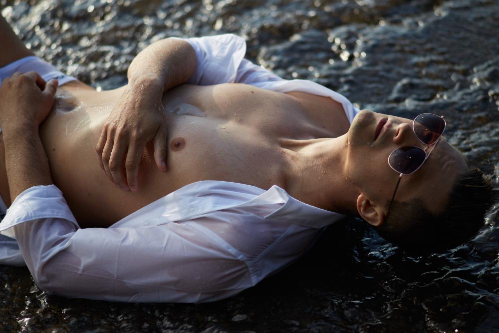 Man in an unbuttoned white shirt and Ray-Ban sunglasses lying in shallow water, with sunlight highlighting his toned chest and relaxed expression.