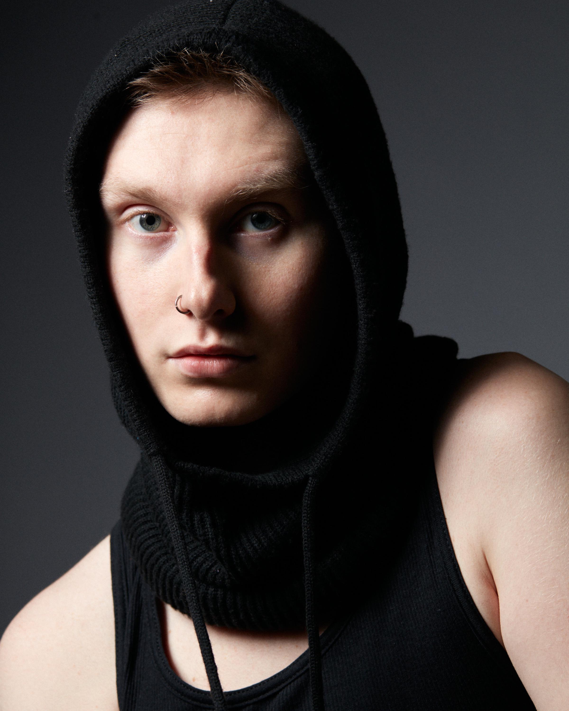 Close-up portrait of a man in a black hooded scarf and tank top, with a nose ring and piercing blue eyes, gazing intensely at the camera.
