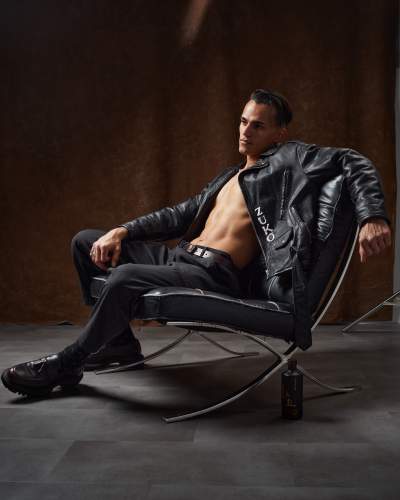 A man reclines in a black leather chair, wearing a black leather jacket and pants, with a bottle of cologne on the floor nearby, set against a dark, textured background.