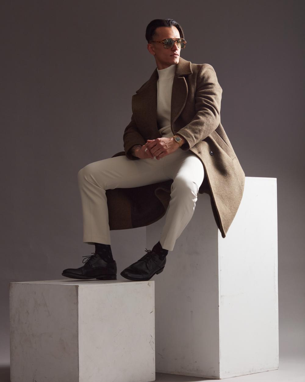 Man wearing a brown overcoat, white turtleneck, and cream trousers sitting stylishly on a white pedestal, accessorized with Gucci unglasses and a green-faced wristwatch, striking a confident pose.