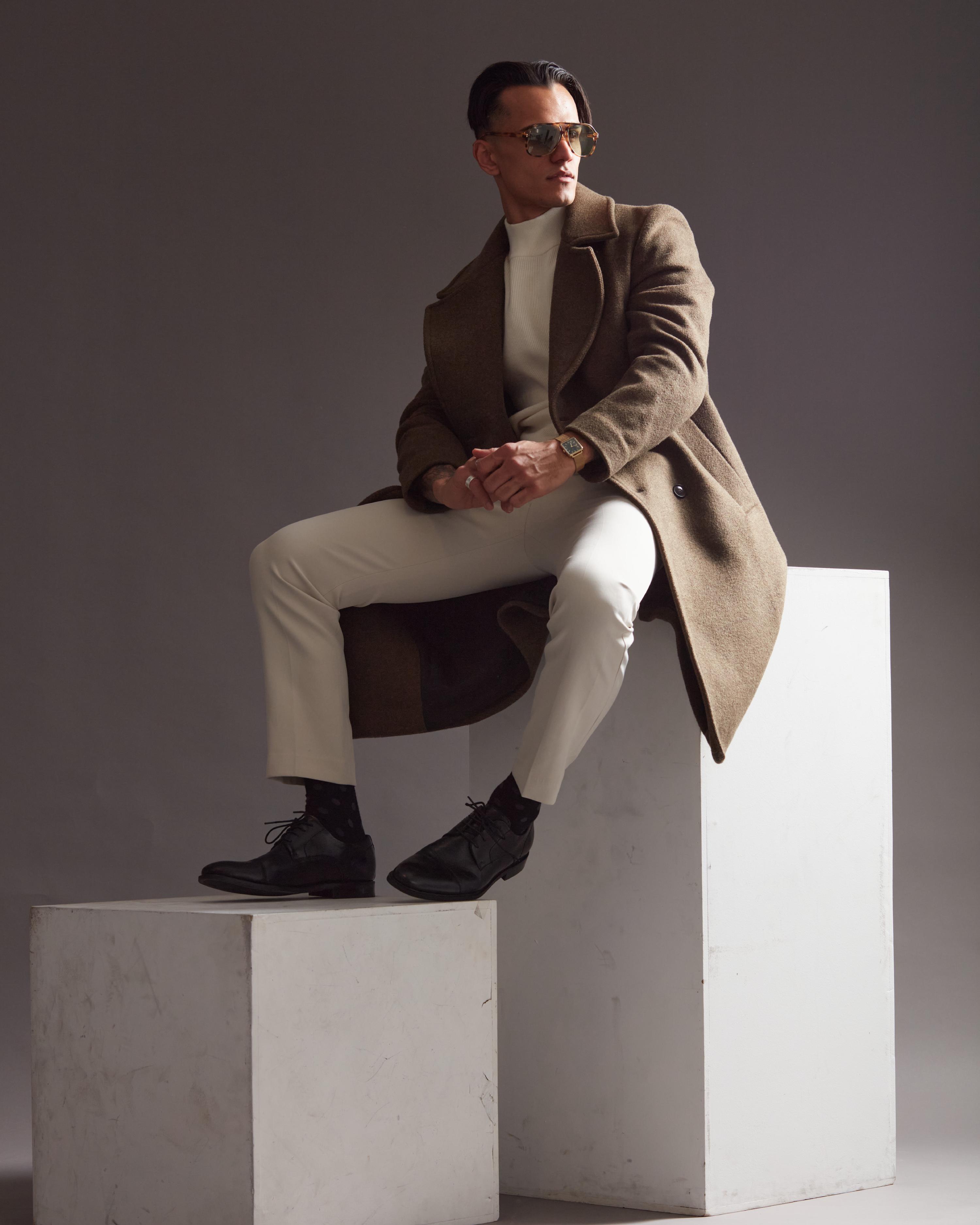 Man wearing a brown overcoat, white turtleneck, and cream trousers sitting stylishly on a white pedestal, accessorized with Gucci unglasses and a green-faced wristwatch, striking a confident pose.
