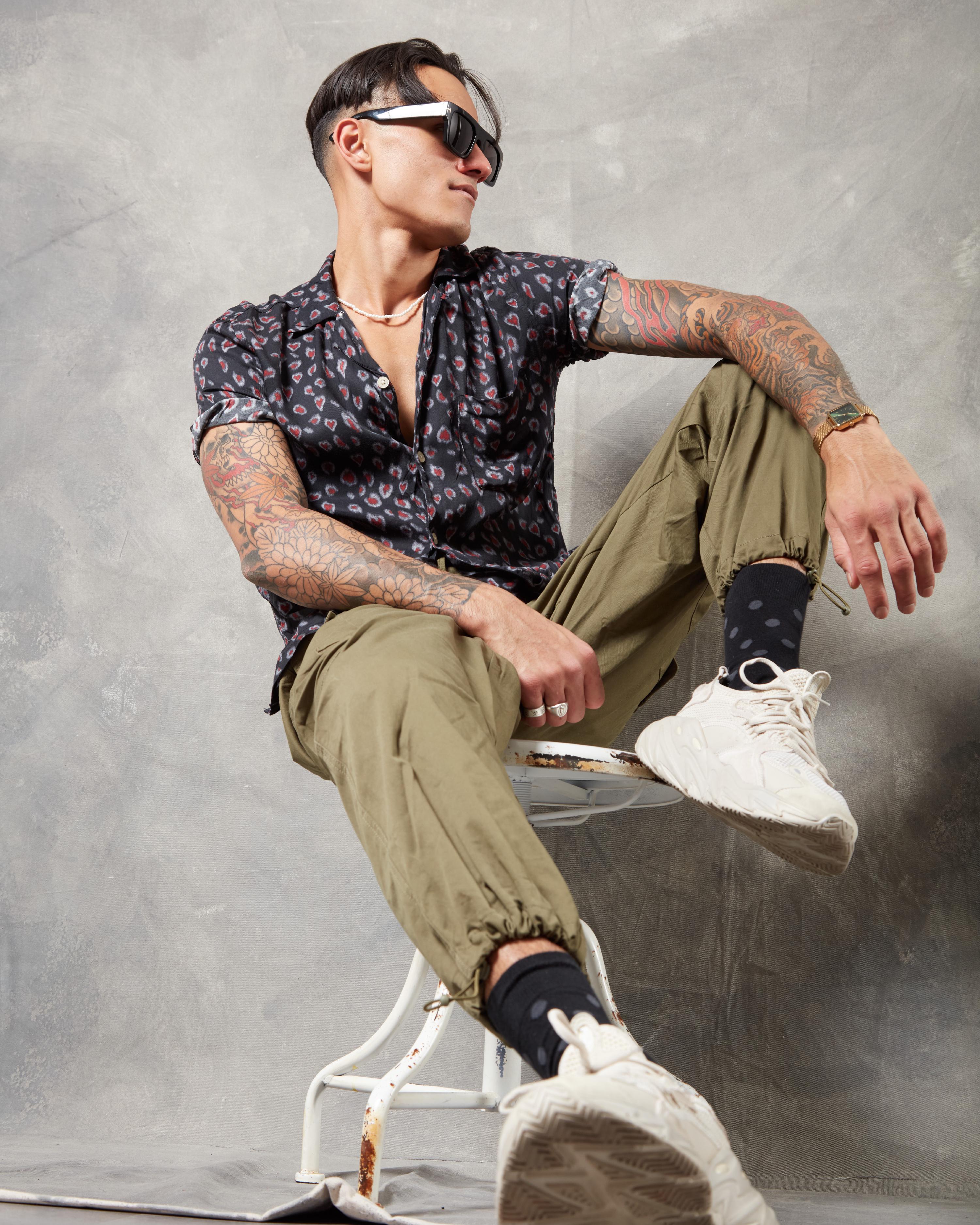 Tattooed man in a patterned short-sleeve shirt, khaki joggers, and white sneakers posing on a vintage chair, accessorized with sunglasses and a gold wristwatch against a textured gray backdrop.
