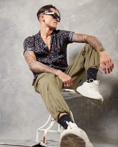 Tattooed man in a patterned short-sleeve shirt, khaki joggers, and white sneakers posing on a vintage chair, accessorized with sunglasses and a gold wristwatch against a Gravity handpainted textured gray backdrop.