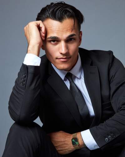 Man in a sharp black suit, white shirt, and black tie posing confidently with his hand on his head, wearing a stylish green-faced wristwatch, and gazing directly at the camera.