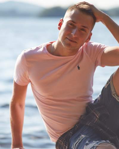 Man in a pink Ralph Lauren t-shirt and denim shorts sitting by a lake, running his hand through his hair and smiling softly in warm sunlight.