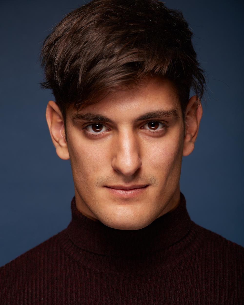Close-up portrait of a man with dark brown hair, wearing a burgundy ribbed turtleneck sweater, gazing confidently into the camera against a dark blue background.