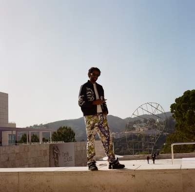 Man in a black jacket with white patterns, graphic print pants, and sunglasses standing confidently at the Getty Center, with hills, a sculpture, and clear blue sky in the background.