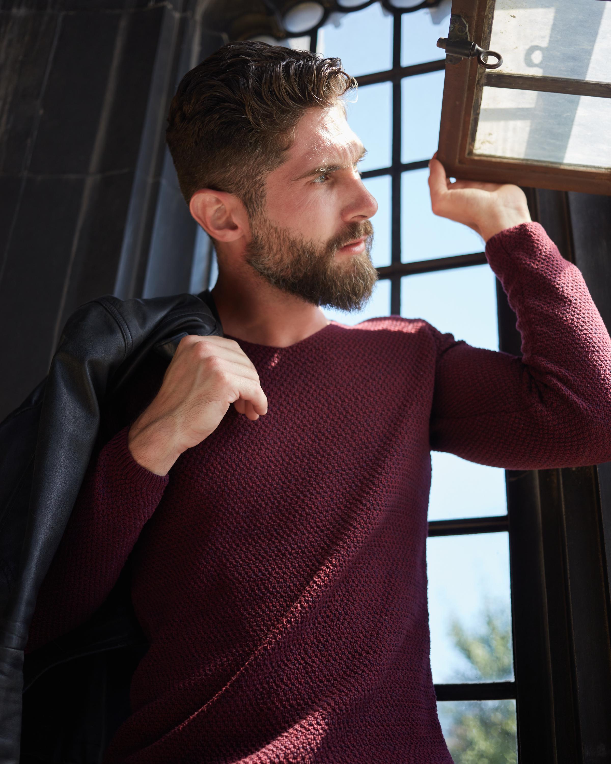 Man with a well-groomed beard wearing a textured burgundy sweater, holding a black leather jacket over his shoulder while opening a window with natural sunlight streaming in.