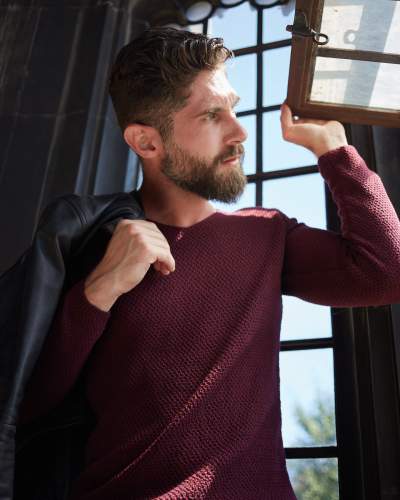 Man with a well-groomed beard wearing a textured burgundy sweater, holding a black leather jacket over his shoulder while opening a window with natural sunlight streaming in.