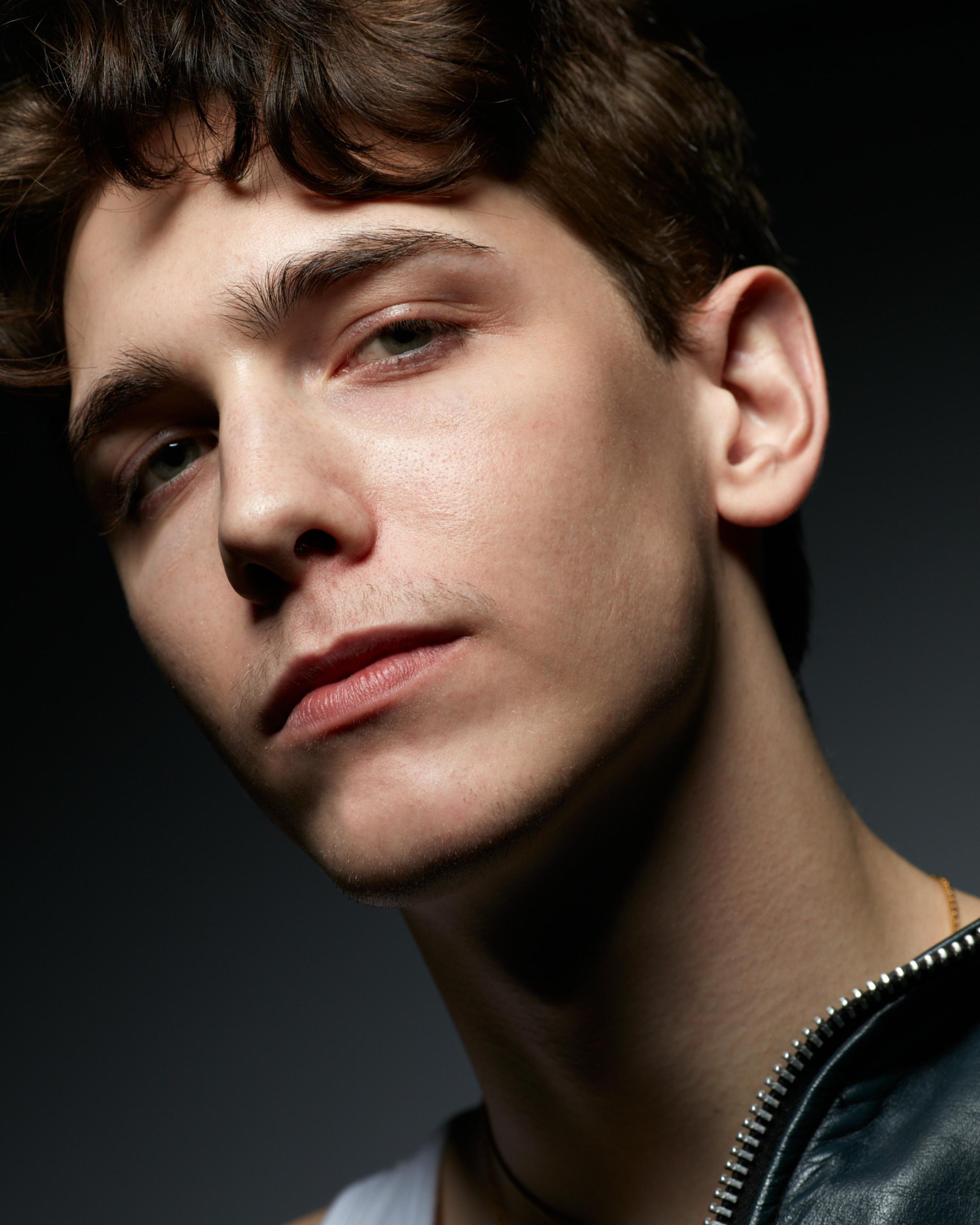 Close-up portrait of a young man with tousled brown hair and a confident gaze, wearing a black leather college jacket and a white tank top against a dark blue background.