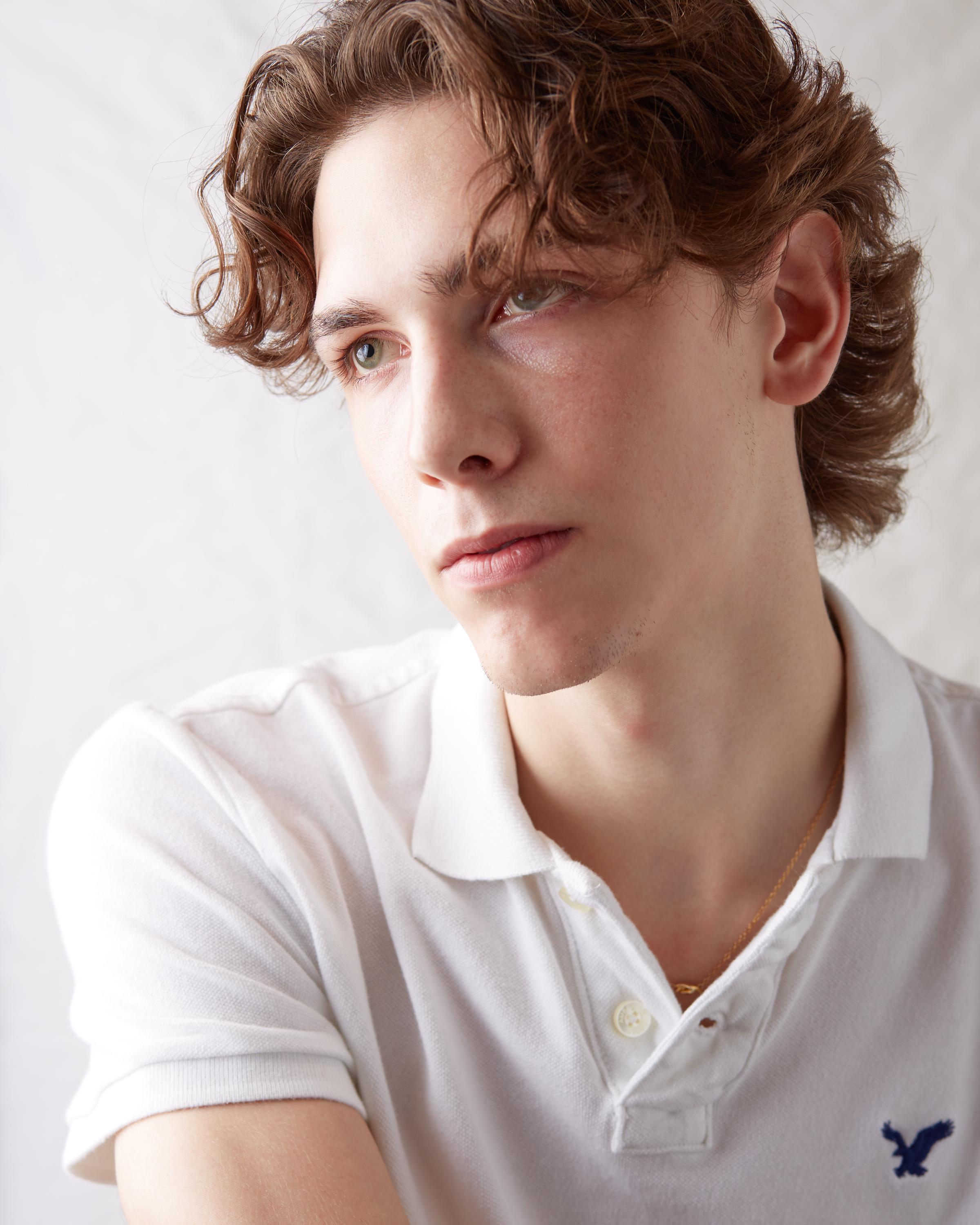 Portrait of a young man with wavy brown hair wearing a white American Eagle polo shirt, gazing thoughtfully to the side against a light background.