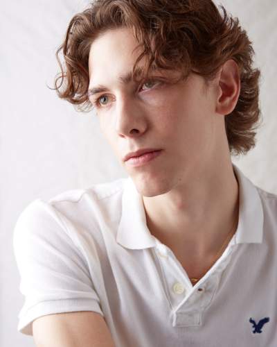 Portrait of a young man with wavy brown hair wearing a white American Eagle polo shirt, gazing thoughtfully to the side against a light background.