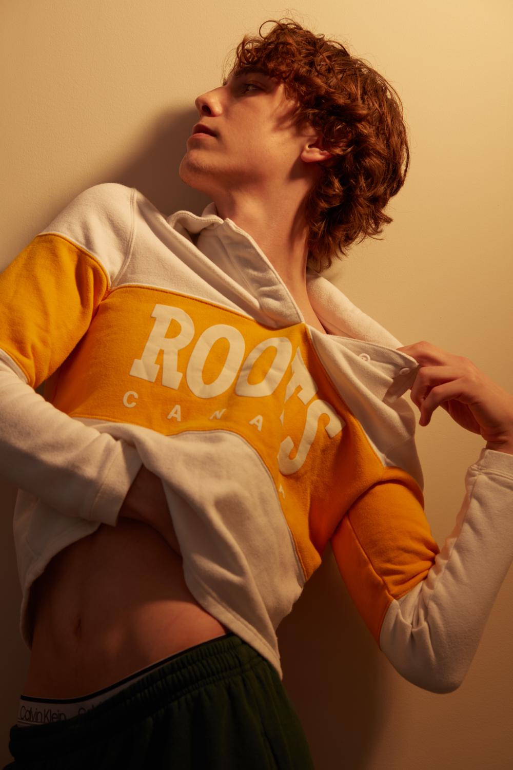 Young man with curly brown hair wearing an orange and white Roots Canada hoodie, lifting it to reveal Calvin Klein waistband, posing against a warm-toned background.