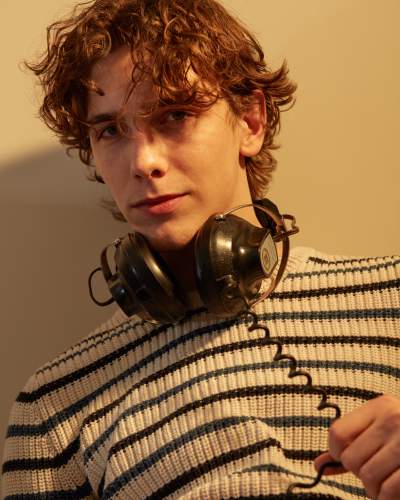 Young man with curly brown hair wearing a striped American Eagle sweater and vintage Koss headphones around his neck, holding the coiled headphone cable against a warm-toned background.