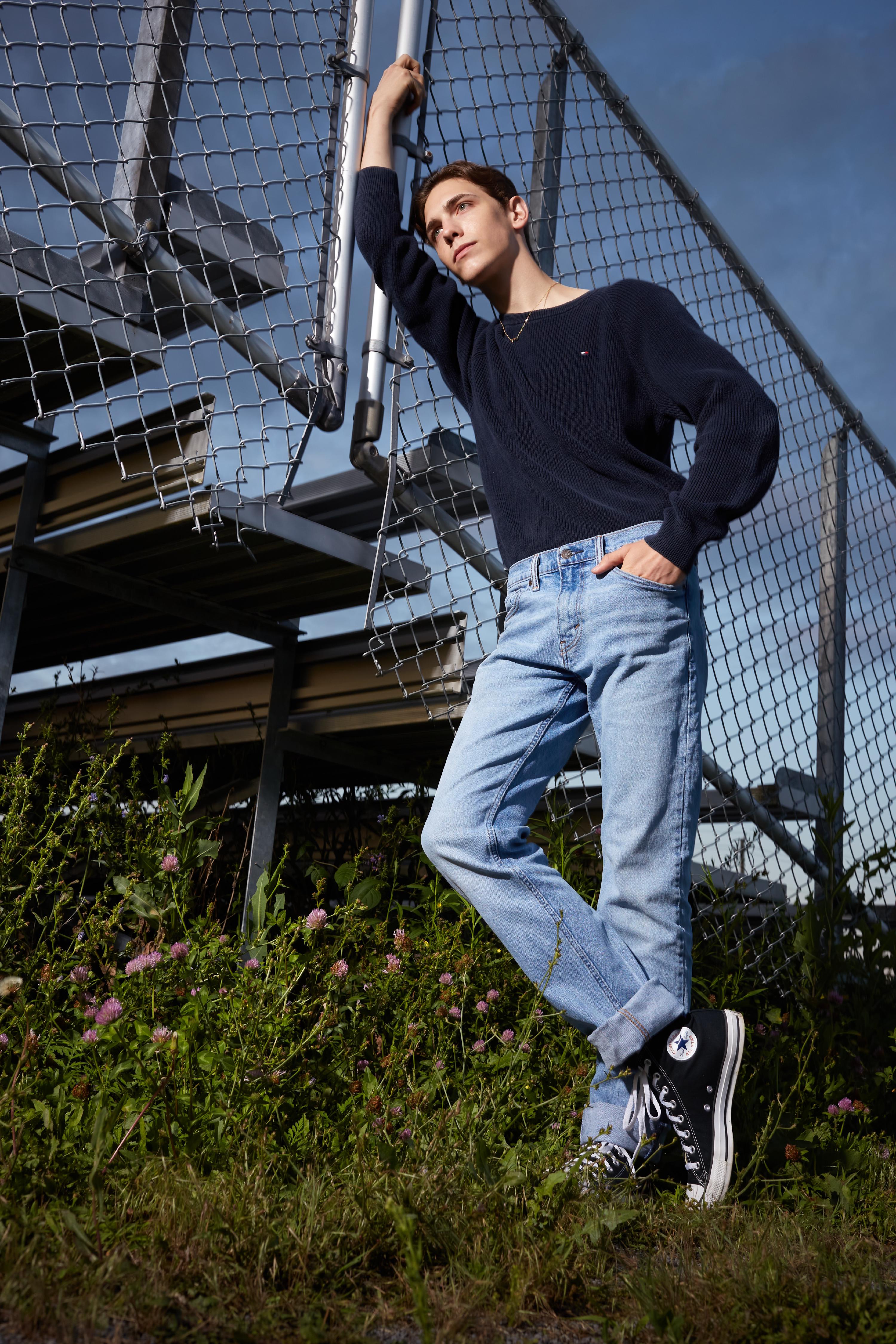 Man in a navy blue Tommy Hilfiger sweater, light blue jeans, and black Converse sneakers leaning casually against a chain-link fence in Bayridge High School, with plants and industrial structures in the background.