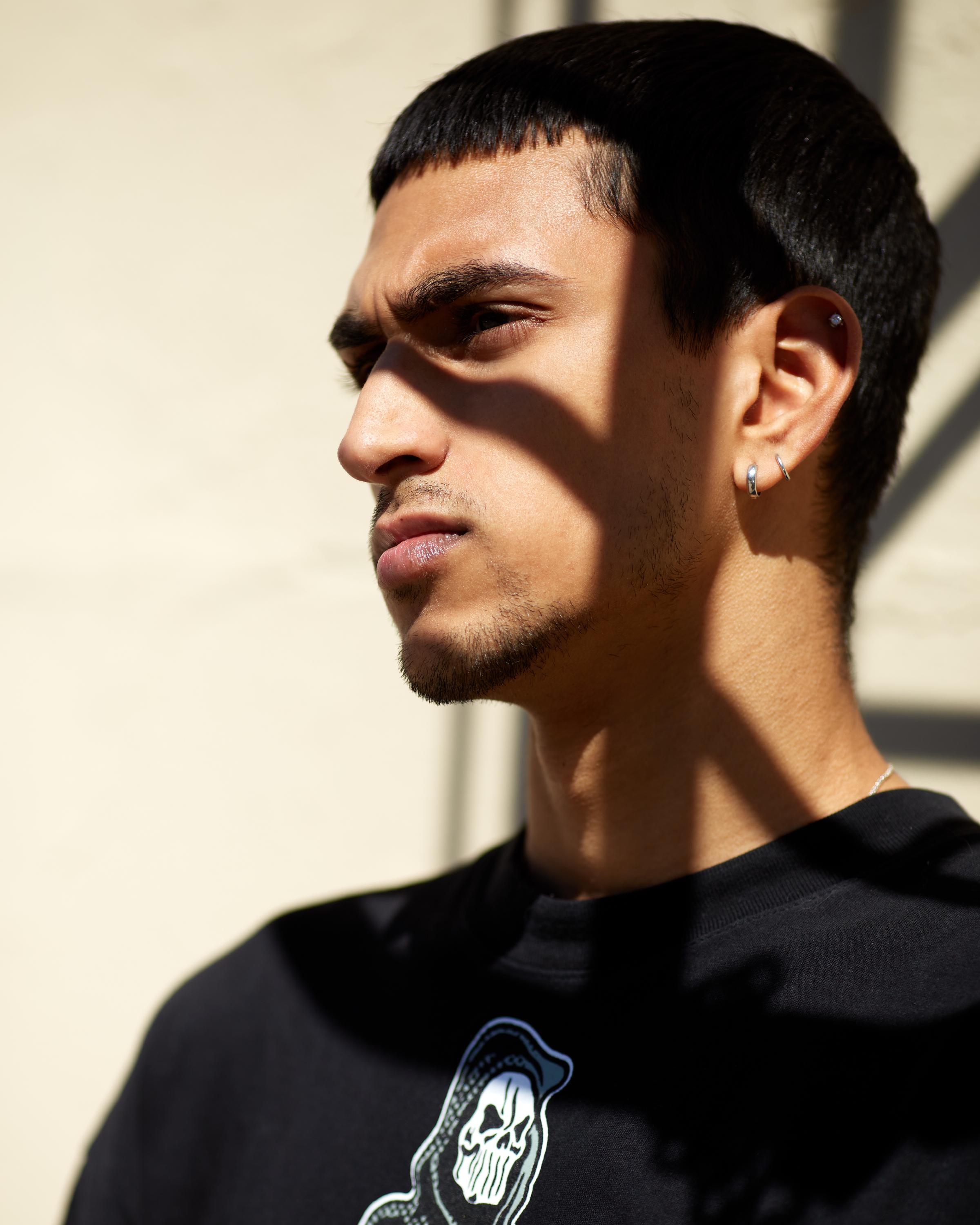 Close-up portrait of a young man with a sharp haircut, wearing small silver hoop earrings and a black graphic t-shirt, with dramatic shadows cast across his face against a neutral background.