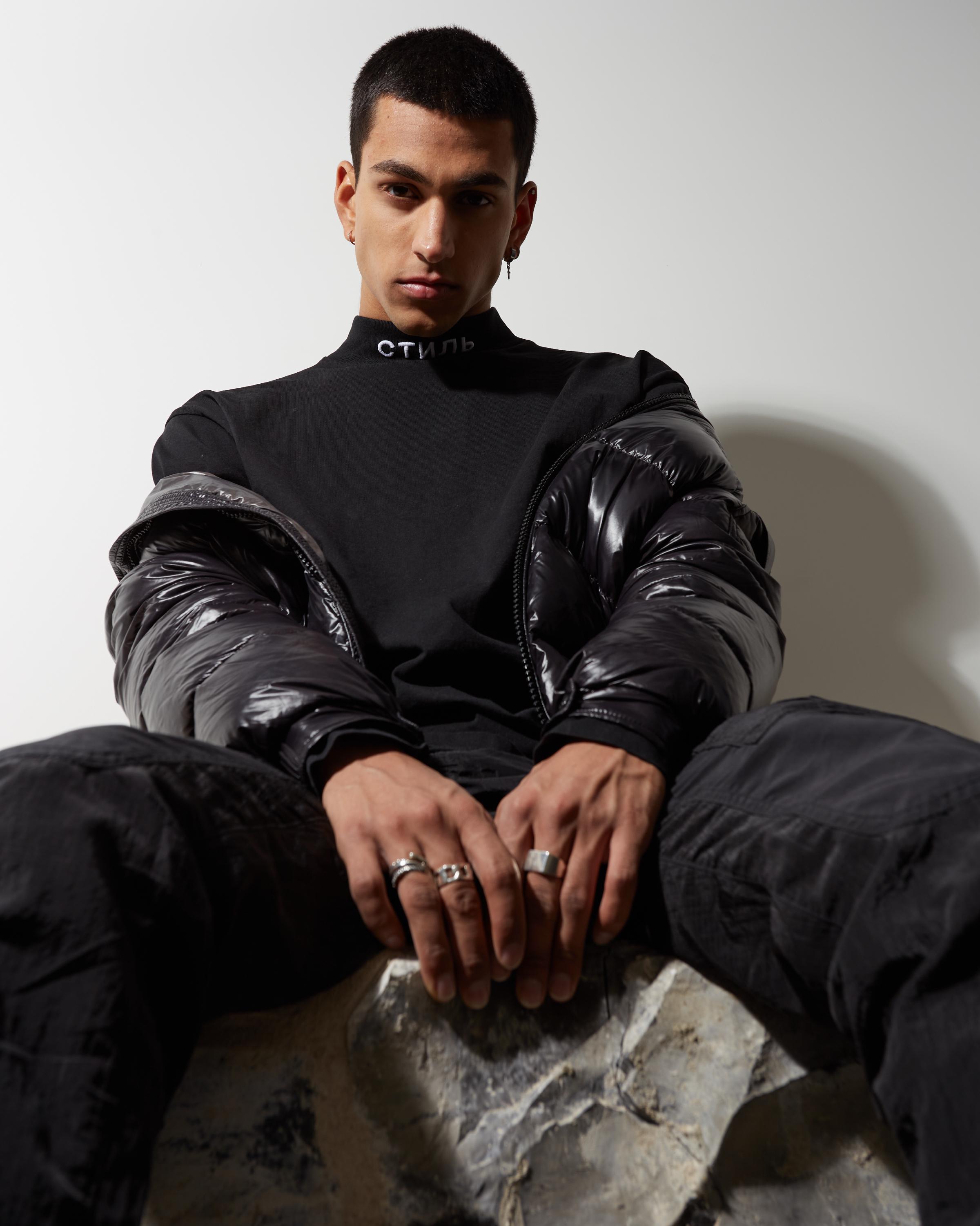 Young man in a black Heron Preston turtleneck with 'CTNMB' Cyrillic text, shiny black puffer jacket, and silver rings, sitting confidently against a neutral background.