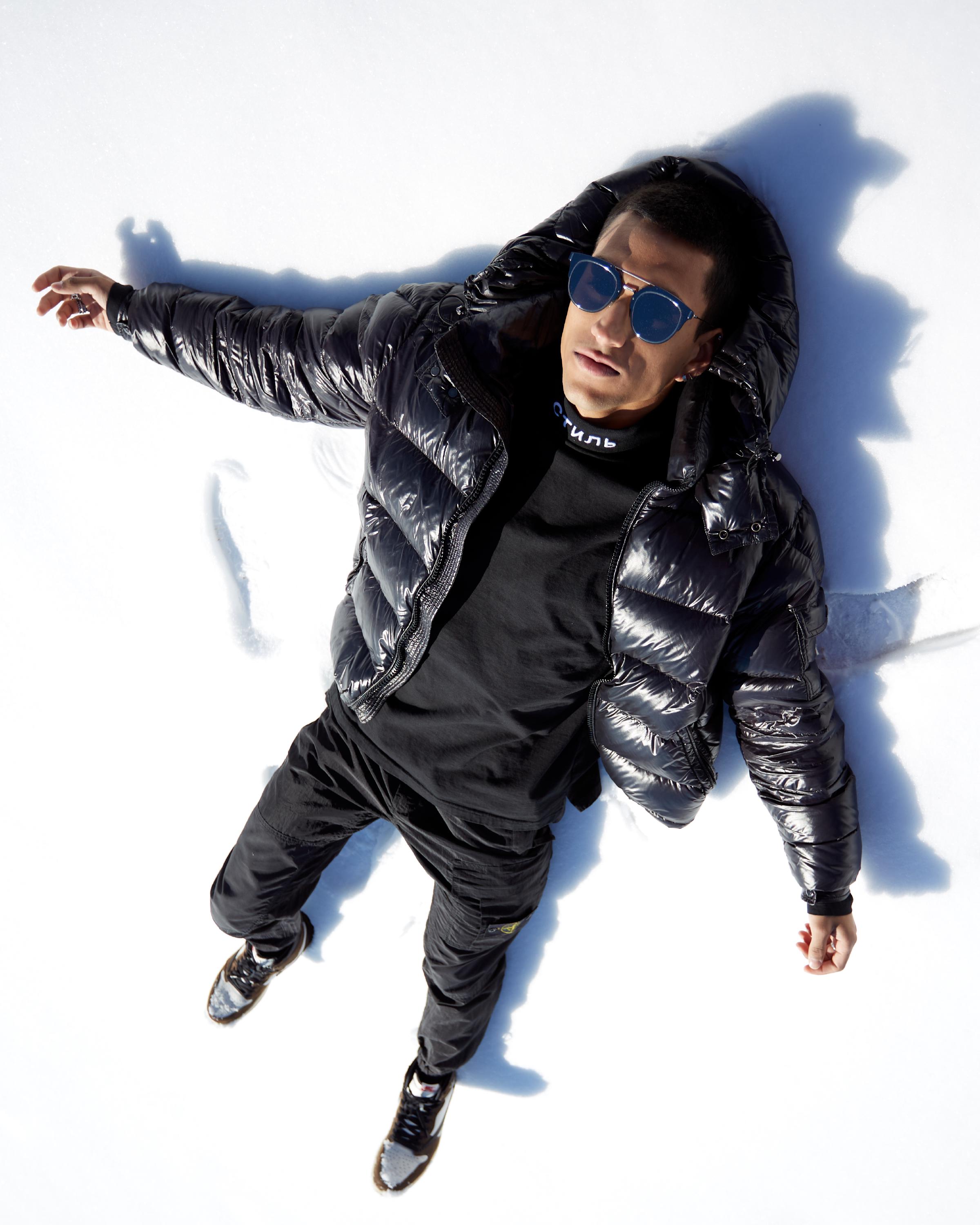 Man lying in the snow wearing a black Heron Preston turtleneck with 'CTNMB' text, shiny black puffer jacket, black cargo pants, and round blue sunglasses, casting a distinct shadow.