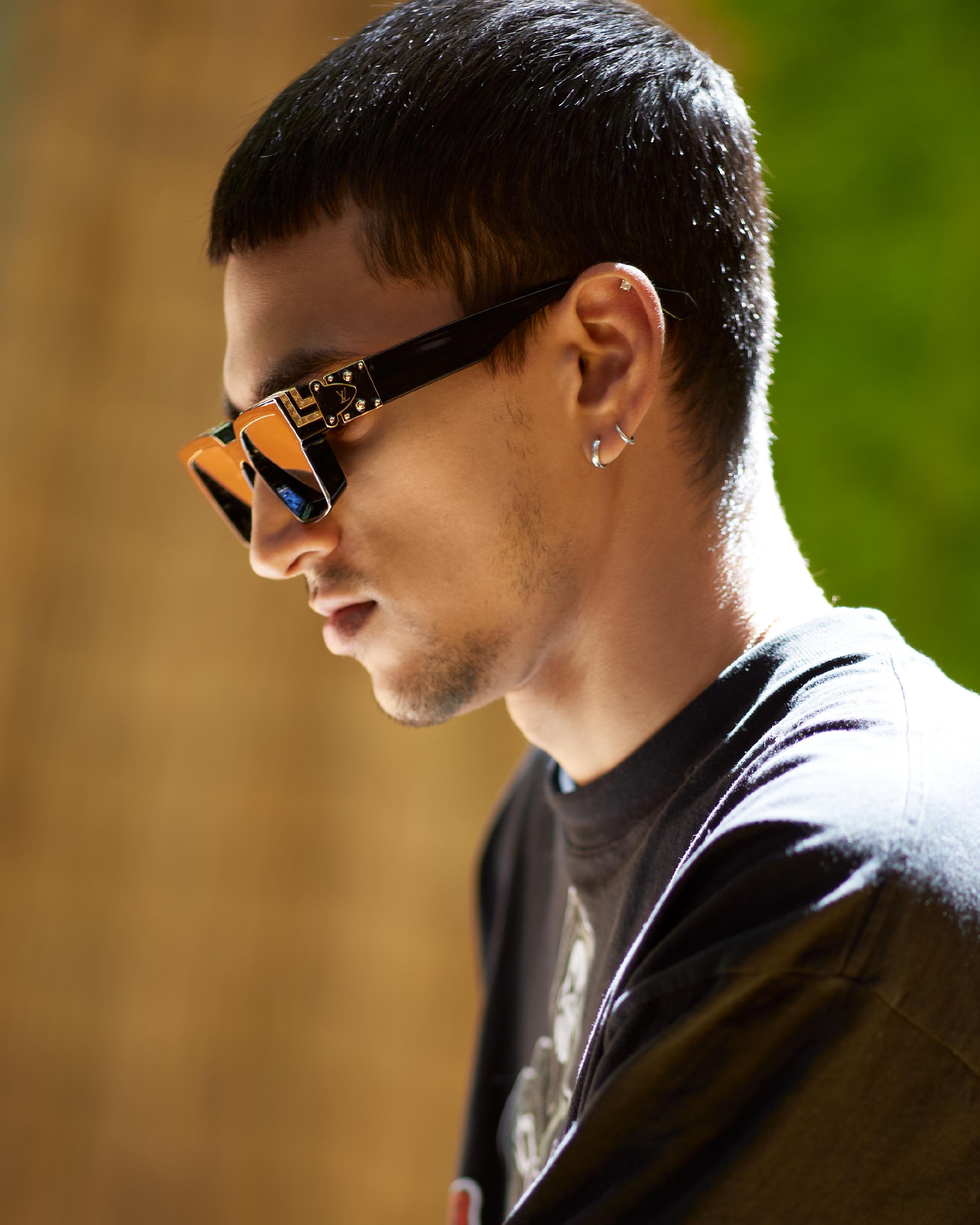 Young man wearing black Louis Vuitton sunglasses and a black graphic t-shirt, with a silver hoop earring, posing against a blurred warm-toned background.
