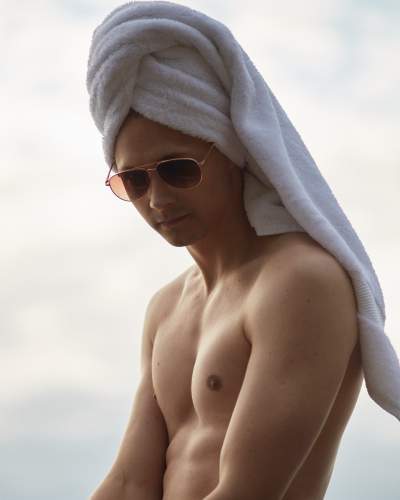 Shirtless man with a white towel wrapped around his head and wearing aviator sunglasses, posing against a soft, neutral sky background.