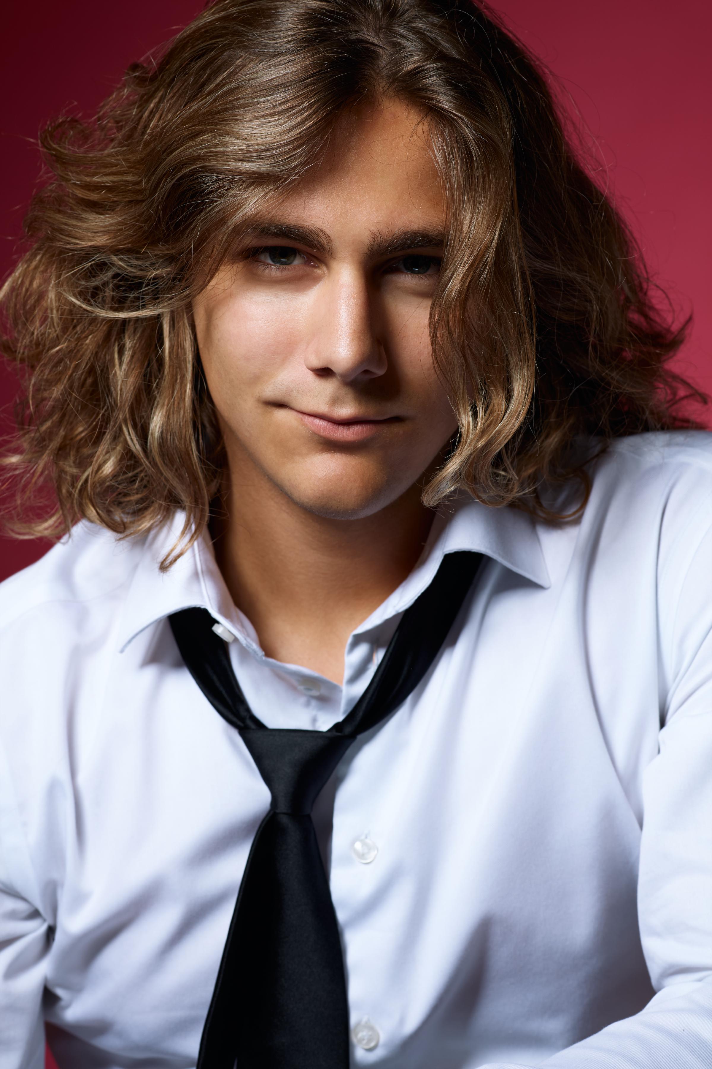 Studio headshot portrait of a young man with long wavy hair wearing a white dress shirt and loosened black tie, subtle smile and direct eye contact on a deep magenta backdrop