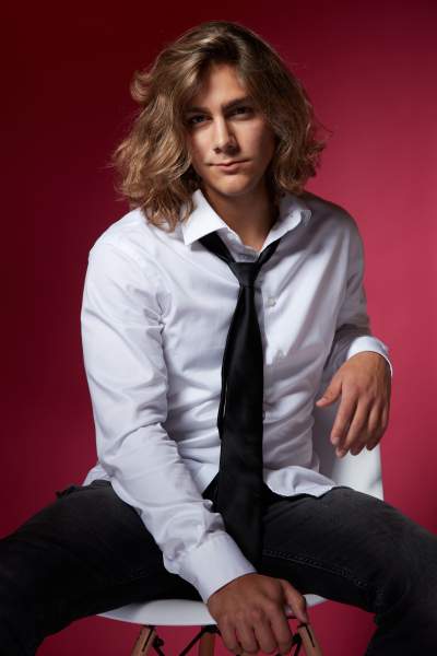 Young man with long wavy hair wearing a white dress shirt, black tie, and dark jeans, seated casually on a white chair against a vibrant red background