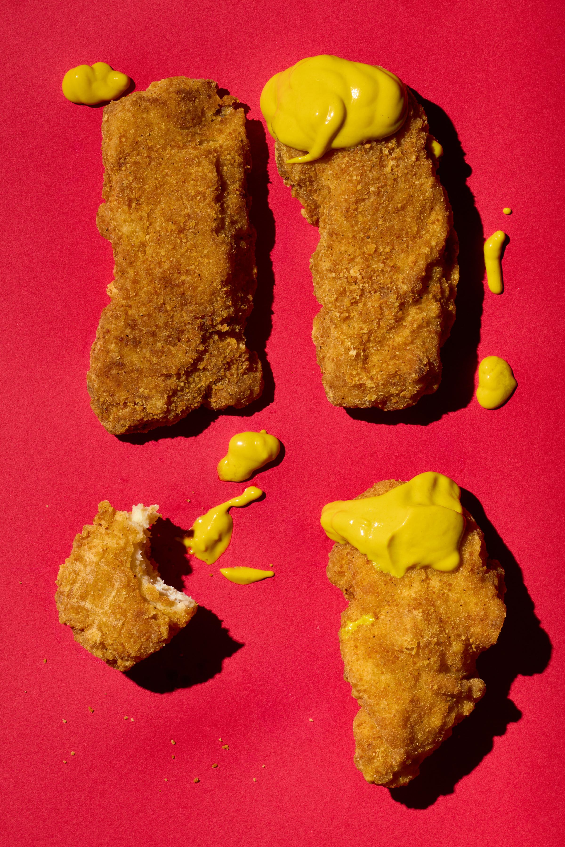 Golden brown chicken nuggets with dollops of yellow mustard, arranged on a bold red background with scattered mustard drips.