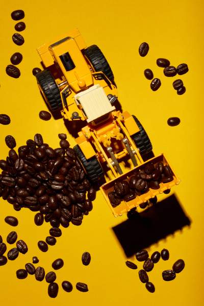 Top-down view of a yellow toy bulldozer scooping up dark roasted coffee beans scattered on a bright yellow background.