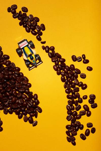 Overhead view of a yellow toy Renault Formula 1 race car driving through scattered dark roasted coffee beans on a vibrant orange background.