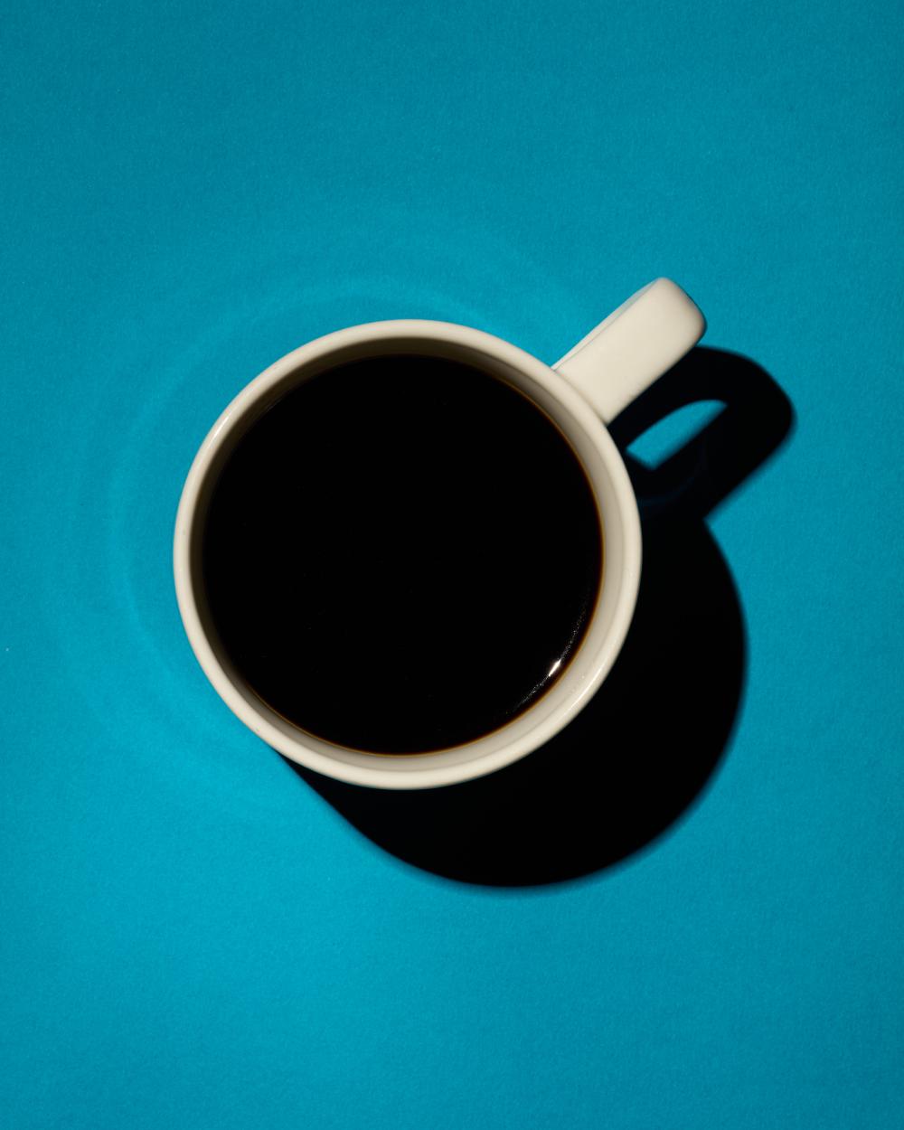 Top-down view of a white coffee mug filled with black coffee, casting a strong shadow on a vibrant blue background.