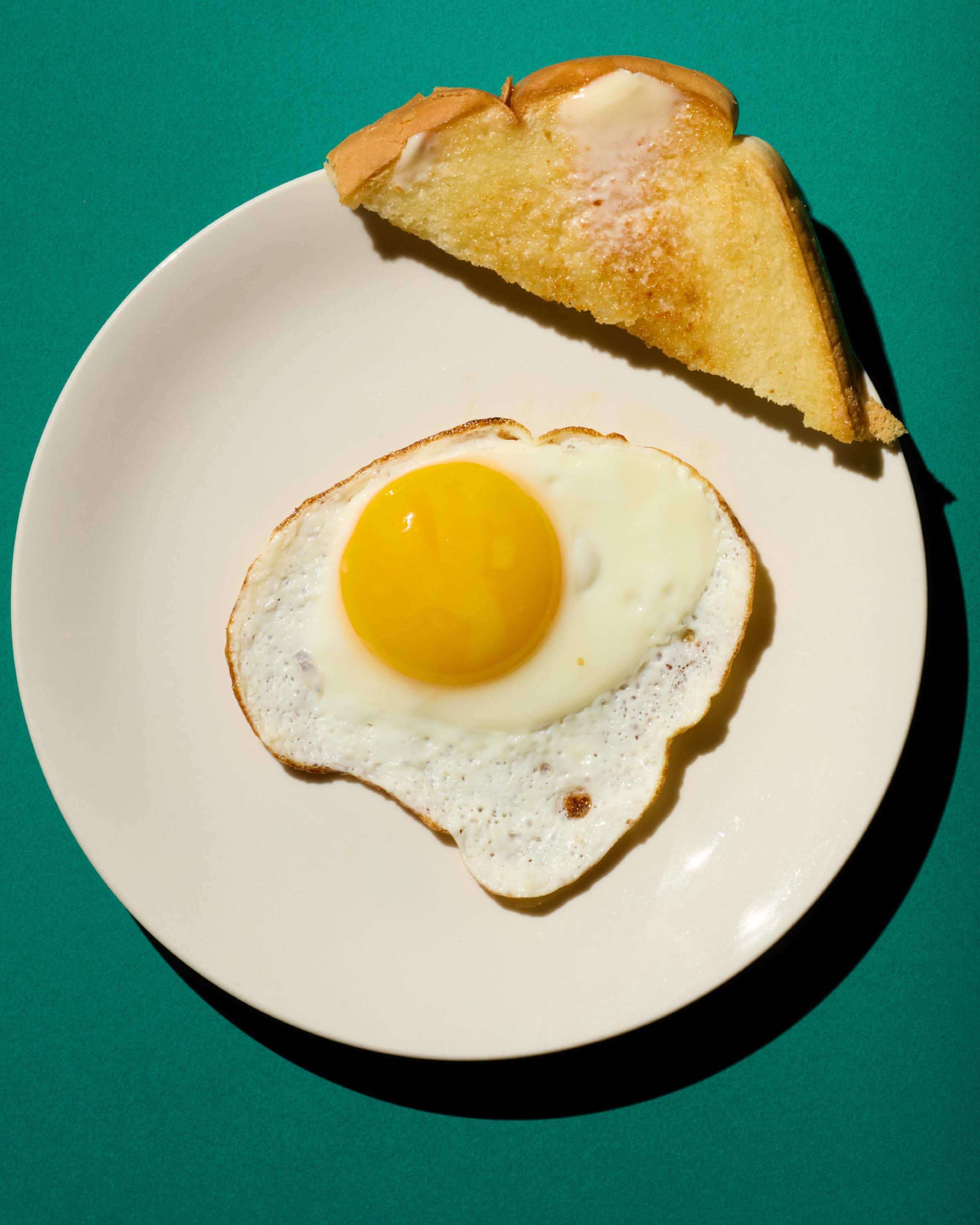 Overhead view of a sunny-side-up fried egg with a vibrant yellow yolk next to a golden-brown buttered toast slice, served on a white plate against a teal background.