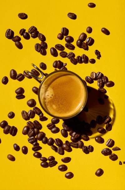 Top-down view of a freshly brewed espresso in a glass cup surrounded by scattered roasted coffee beans on a vibrant yellow background, casting rich shadows and creating a bold, textured composition.