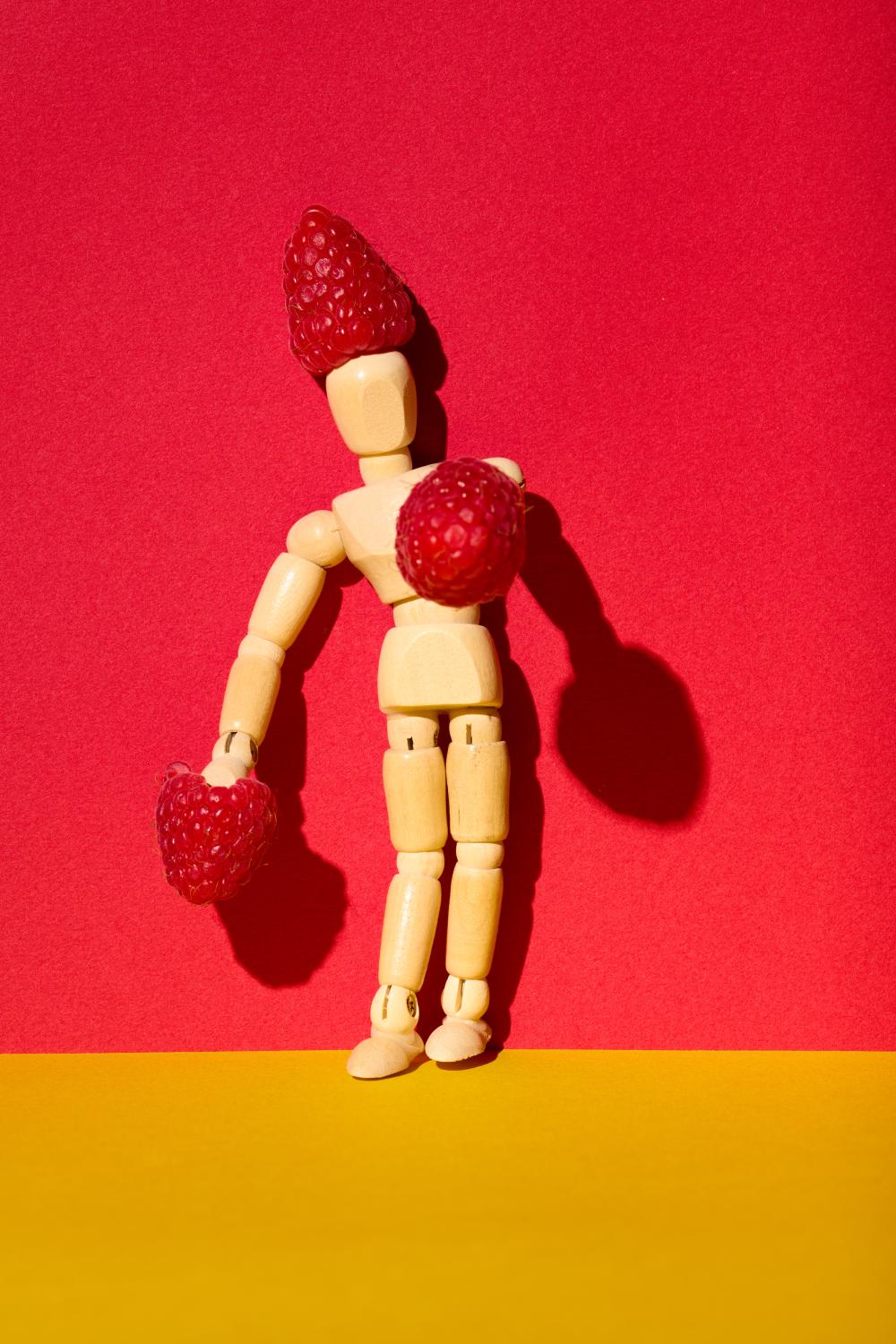 A wooden artist mannequin posed against a vibrant red background, creatively adorned with fresh raspberries as a whimsical hat and gloves. The bold yellow surface adds contrast, enhancing the playful and surreal composition.