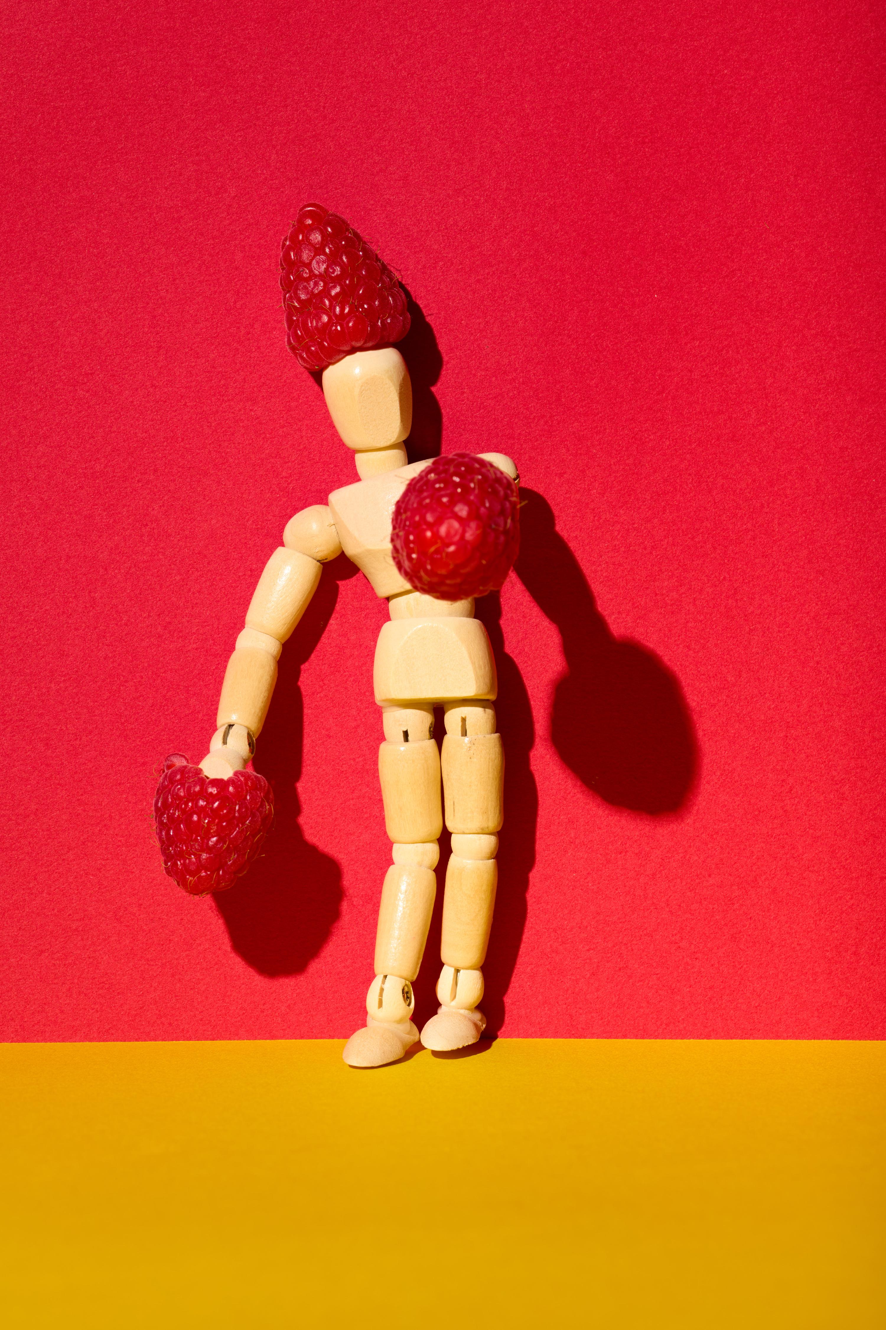 A wooden artist mannequin posed against a vibrant red background, creatively adorned with fresh raspberries as a whimsical hat and gloves. The bold yellow surface adds contrast, enhancing the playful and surreal composition.