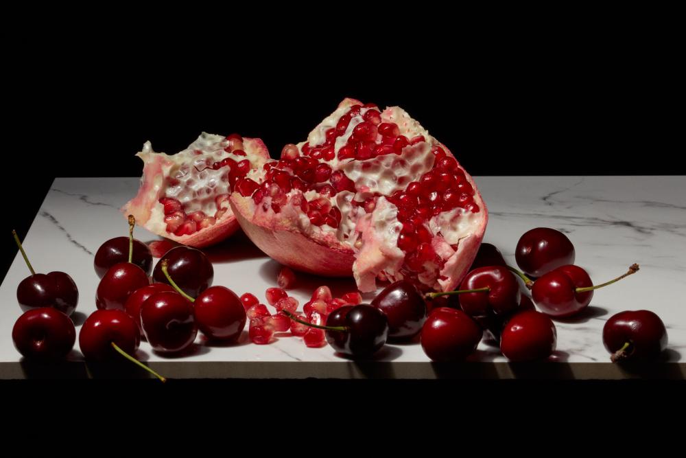 Pomegranate halves with glistening red seeds surrounded by fresh cherries on a marble surface that are about to roll off the edge.