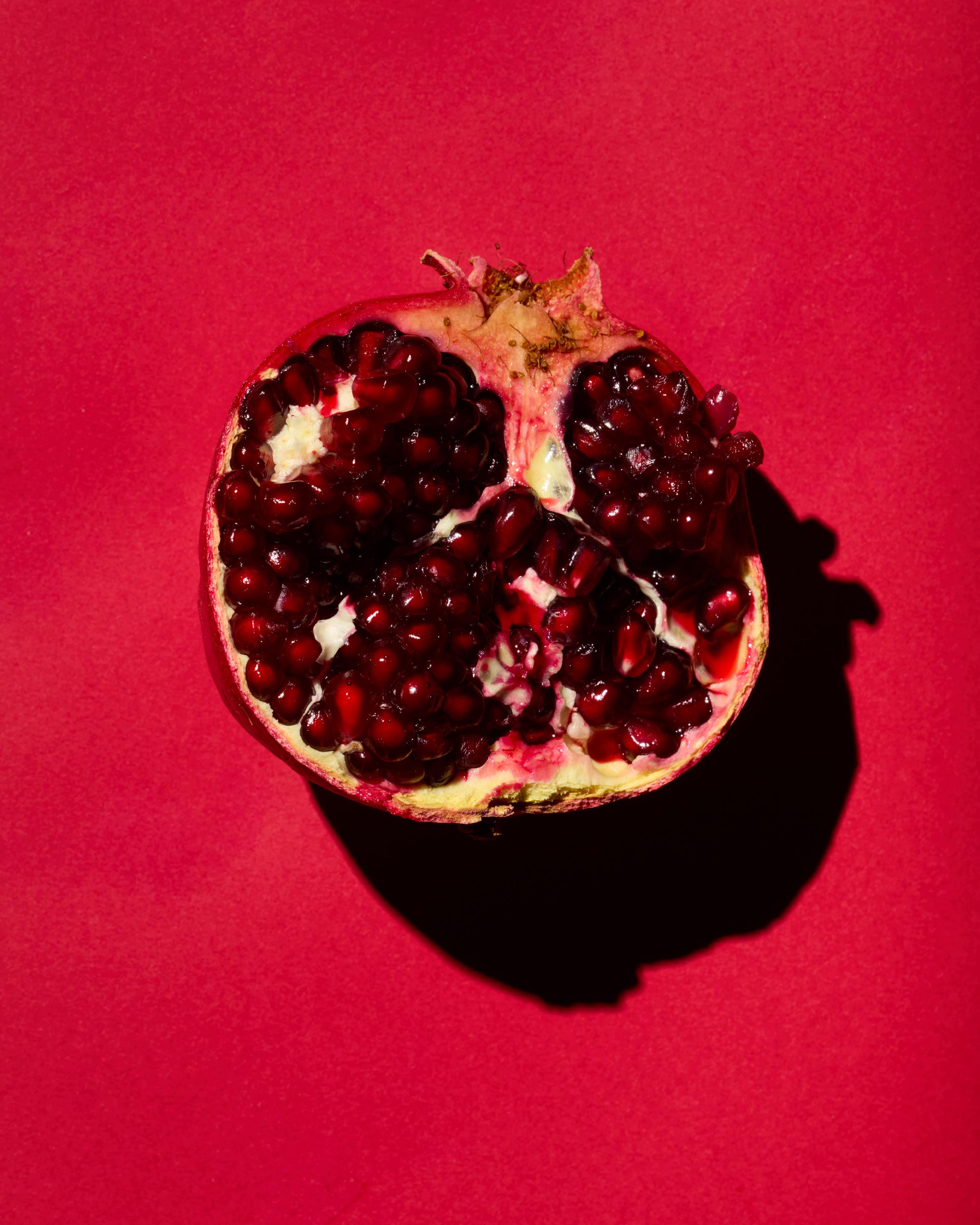 A halved pomegranate with deep red seeds on a bold red background, casting a dramatic shadow.