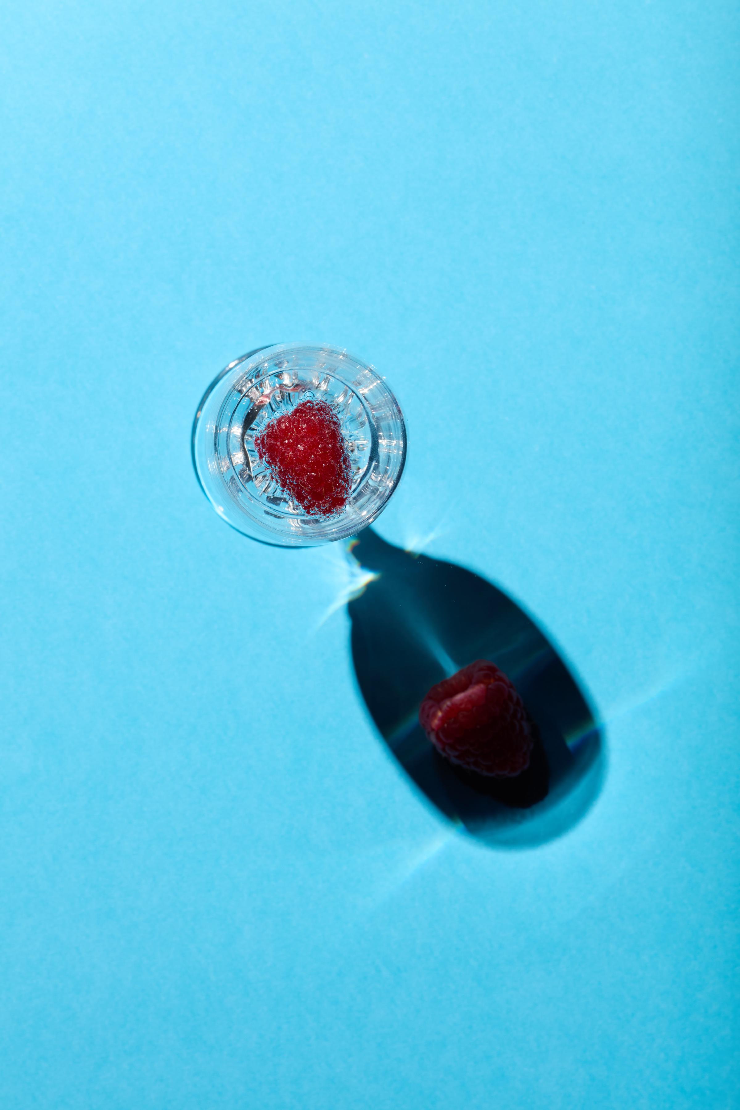 A raspberry submerged in a glass of sparkling water, casting a shadow on a vibrant blue background.