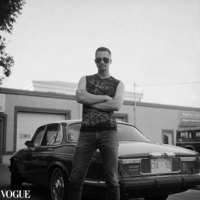 Black and white photo of a man wearing Ray Ban aviator sunglasses, a sleeveless graphic shirt, and jeans, standing confidently with crossed arms in front of a vintage Jaguar car, with Vogue branding in the corner.