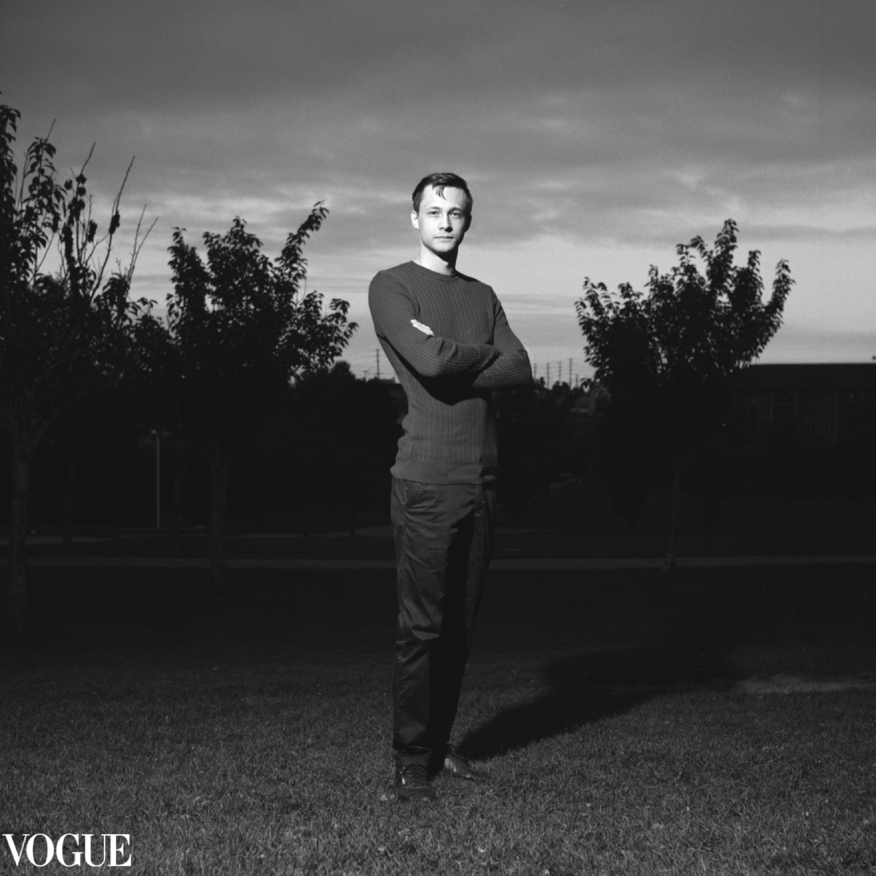 Black and white portrait of a man standing in a park with crossed arms, wearing a textured sweater and dark pants, under a dramatic sky, with Vogue branding in the corner.